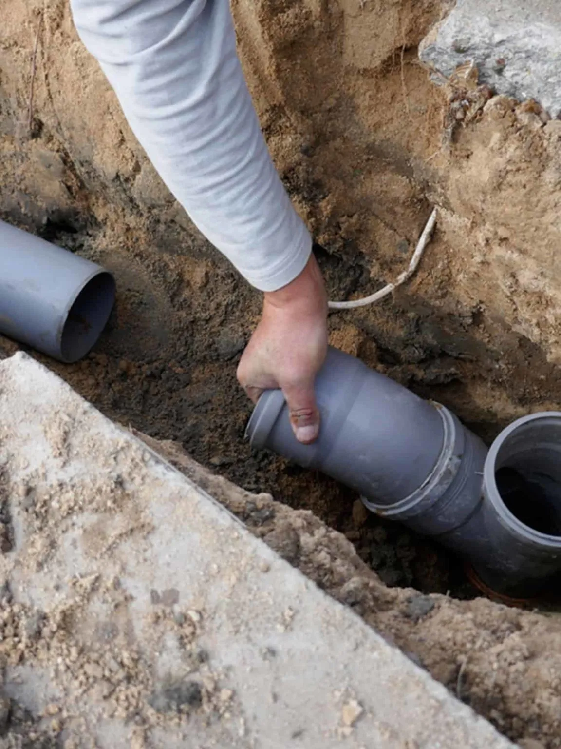 Engineer fitting a new grey plastic drain pipe into a joint in an open trench