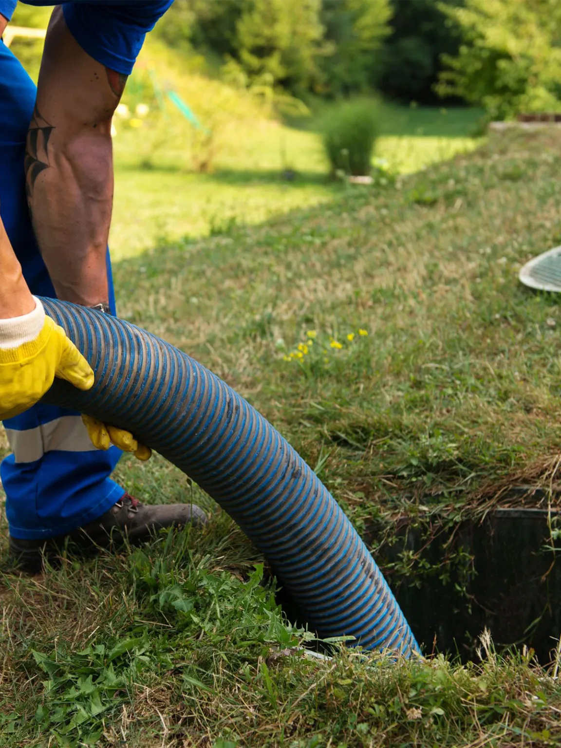 Engineer in blue overalls feeding a tanker suction hose into an open septic tank chamber in a garden
